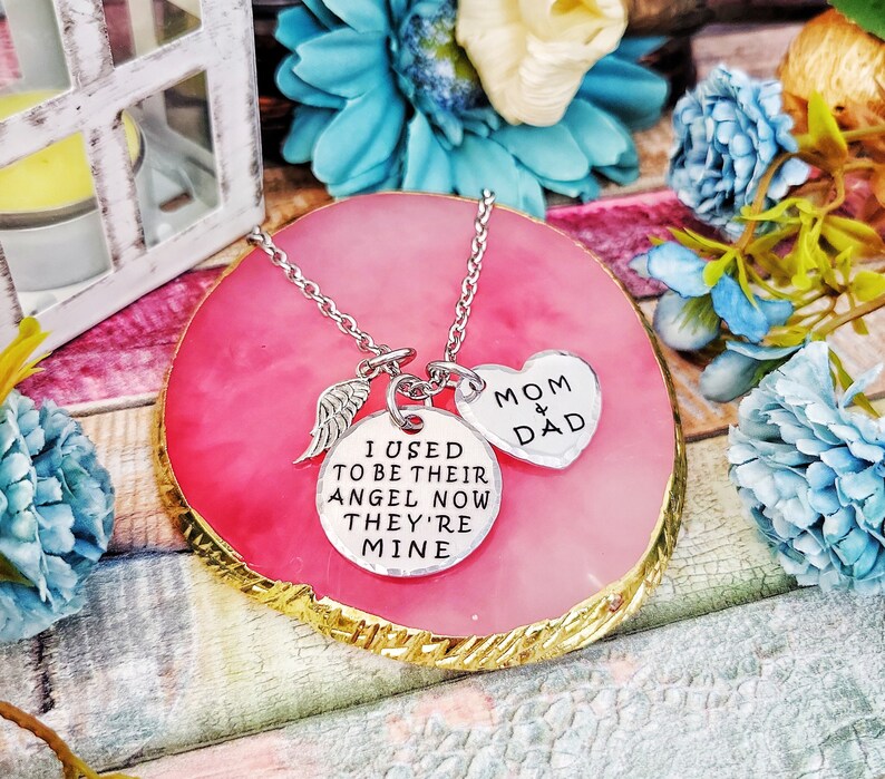 May include: Silver necklace with three charms: a wing, a round charm with the text "I used to be their angel now they're mine", and a heart charm with the text "Mom & Dad".