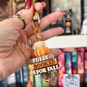 May include: A keychain featuring a gold-toned metal ring, two stacked miniature pumpkins in cream and orange, and three rectangular beads resembling book spines. The beads are inscribed with the text FULLY BOOKED FOR FALL in white and black.