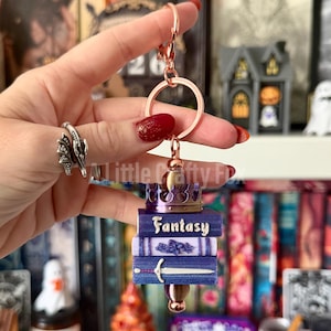 May include: A rose gold keychain featuring a stack of three miniature books in shades of purple and blue. The top book is labeled Fantasy, the middle has a textured spine, and the bottom features a silver sword. A small crown sits on top.