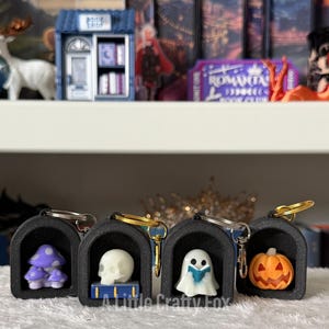 May include: Four black arched keychains featuring miniature figures: a purple mushroom, a white skull on a blue book, a white ghost reading a blue book, and an orange jack-o'-lantern. Each keychain has a gold or silver metal ring and clasp.