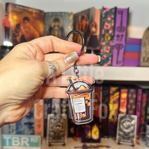 May include: A clear acrylic keychain featuring a cartoon illustration of a coffee cup. The cup is filled with a brown liquid and topped with whipped cream, caramel drizzle, and ice cubes. The text on the cup reads "Order 1 of 1 Name in order 1: Romantasy 2x Ice 2x Caramel Drizzle 1x Whipped Cream Extra Flavor: Pumpkin Spice"