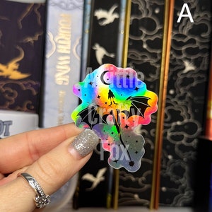 May include: A holographic sticker featuring a black silhouette of a dragon flying through a rainbow-colored sky with stars and a crescent moon. The sticker has the text "A Little Crazy Fox" at the bottom.