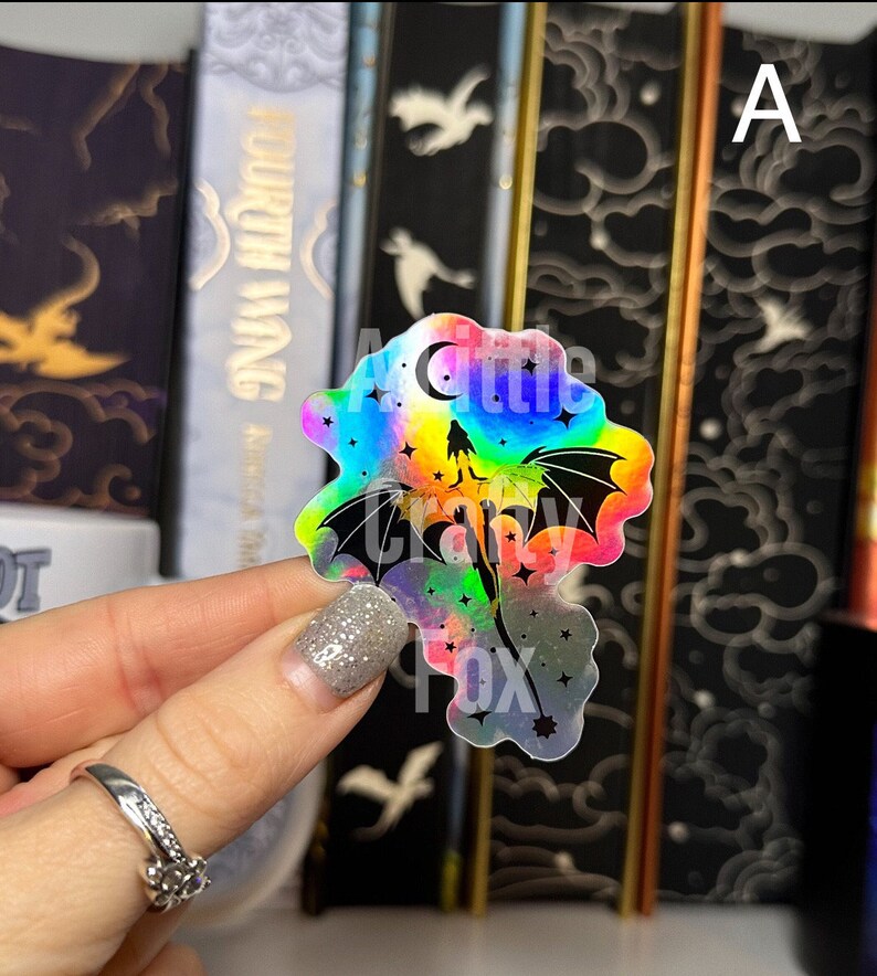 May include: A holographic sticker featuring a black silhouette of a dragon flying through a rainbow-colored sky with stars and a crescent moon. The sticker has the text "A Little Crazy Fox" at the bottom.