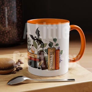 May include: A white ceramic mug with an orange rim and handle. The mug features a black dragon, a vial, a quill pen, and inkwell, and five books with titles including "Navarre" and "Fourth Wing".