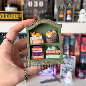 May include: A miniature green bookshelf featuring two shelves filled with small items. The top shelf holds a sleeping orange fox on books and a basket of mushrooms. The bottom shelf contains stacks of purple and orange books and a sleeping creature.