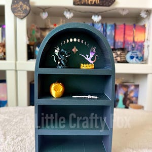 May include: A dark teal miniature bookshelf with three shelves. The top shelf features a black dragon figurine, a purple and pink dragon figurine, and a gold crown. The middle shelf has a gold apple and a silver bracelet. The text "Little Crafty Fox" is visible.