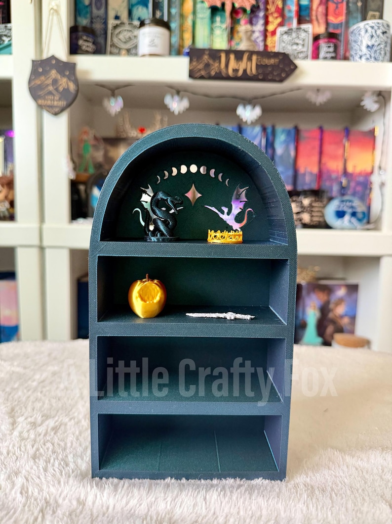 May include: A dark teal miniature bookshelf with three shelves. The top shelf features a black dragon figurine, a purple and pink dragon figurine, and a gold crown. The middle shelf has a gold apple and a silver bracelet. The text "Little Crafty Fox" is visible.