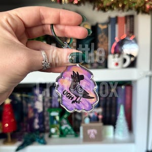 May include: A clear acrylic keychain featuring a cartoon bird with a black hat and the word "UGH" in black text. The bird is set against a purple and orange watercolor background, attached to a green metal ring.