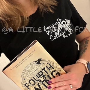 Pu&ograve; includere: Maglietta nera con la scritta "Basgiath War College" e una grafica di un drago. Il libro "Fourth Wing" &egrave; tenuto davanti alla maglietta. La copertina del libro &egrave; color cachi con scritte dorate e un design circolare.