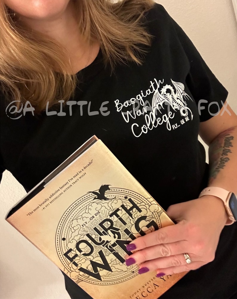 Pu&ograve; includere: Maglietta nera con la scritta "Basgiath War College" e una grafica di un drago. Il libro "Fourth Wing" &egrave; tenuto davanti alla maglietta. La copertina del libro &egrave; color cachi con scritte dorate e un design circolare.