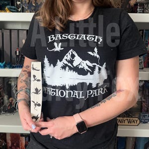 May include: Black t-shirt with a white graphic of mountains, trees, and dragons, with the text "Basgiath National Park." A wooden bookmark displays dragon silhouettes. The theme is fantasy and nature.