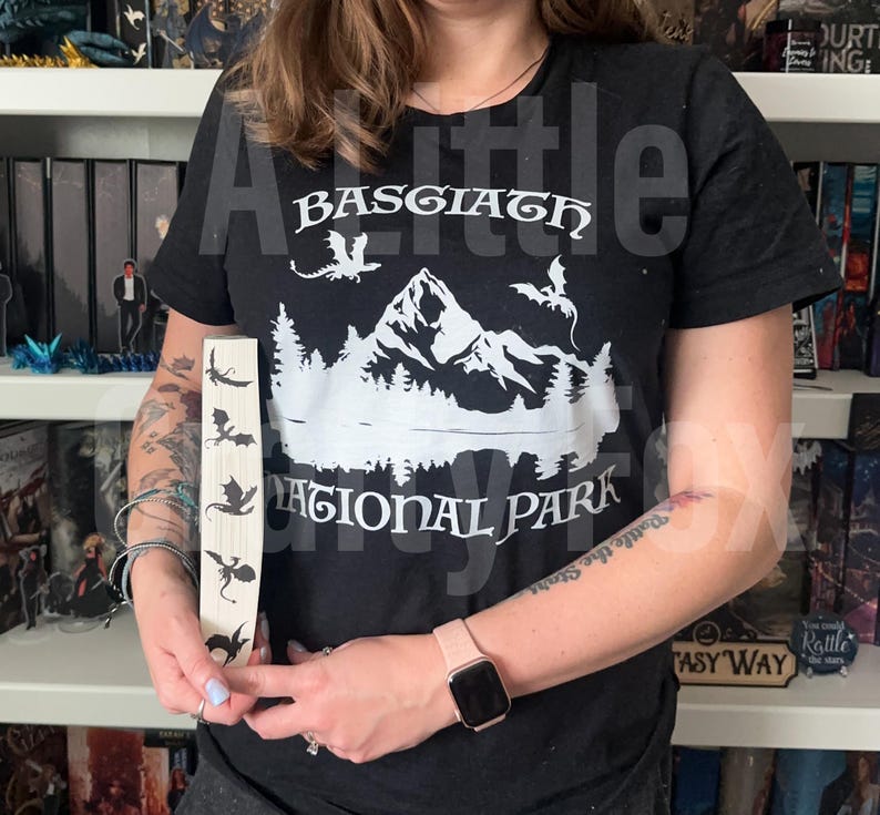 May include: Black t-shirt with a white graphic of mountains, trees, and dragons, with the text "Basgiath National Park." A wooden bookmark displays dragon silhouettes. The theme is fantasy and nature.