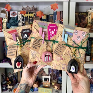 May include: Three rectangular packages wrapped in floral paper, each tied with green twine and featuring a unique keychain. The packages contain stationery items, including sticky notes and bookmarks, with handwritten tags describing book genres.