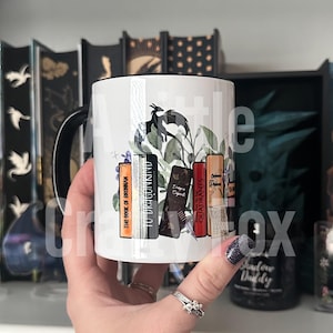 May include: White ceramic mug with a black handle. The mug features a black dragon with green leaves and a stack of books with titles "The Book of Brennan", "To Dragonkind", "Dragon Sigils", "Quadrants", and "Origin of the Phoenix".