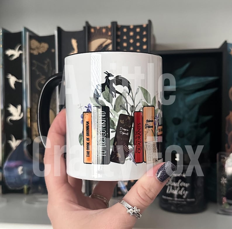 May include: White ceramic mug with a black handle. The mug features a black dragon with green leaves and a stack of books with titles "The Book of Brennan", "To Dragonkind", "Dragon Sigils", "Quadrants", and "Origin of the Phoenix".