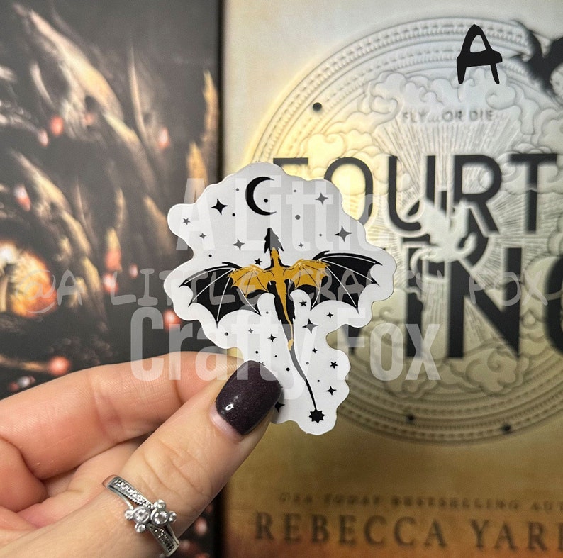 May include: A white sticker with a black and gold dragon flying against a crescent moon and a starry night sky. The sticker is being held by a hand with dark nail polish.