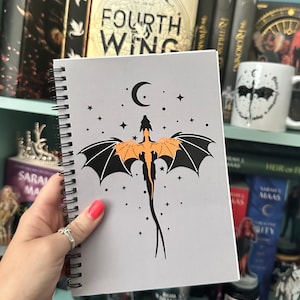 May include: A gray spiral notebook with a black and orange dragon illustration. The dragon is flying with its wings spread and a crescent moon is above it. There are small white stars scattered around the dragon and moon.
