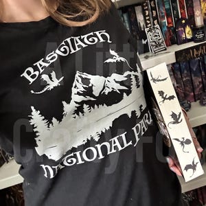 May include: Black t-shirt with white graphic of mountains, trees, and dragons, with the text "Basgiath Regional Park". A wooden bookmark with dragon silhouettes is held next to the shirt.