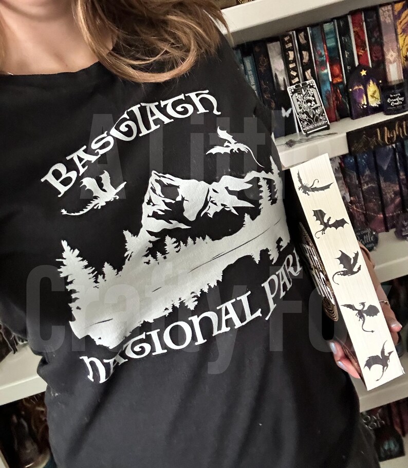 May include: Black t-shirt with white graphic of mountains, trees, and dragons, with the text "Basgiath Regional Park". A wooden bookmark with dragon silhouettes is held next to the shirt.