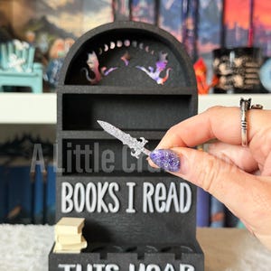 May include: Black wooden bookshelf with the text "BOOKS I READ THIS YEAR". The bookshelf has a decorative top with dragon and star cutouts. A small stack of books sits on the bottom shelf. A silver sword is held in front of the shelf.