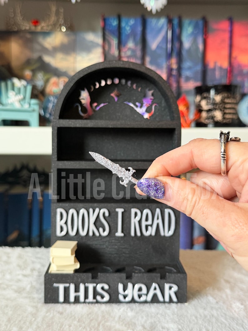 May include: Black wooden bookshelf with the text "BOOKS I READ THIS YEAR". The bookshelf has a decorative top with dragon and star cutouts. A small stack of books sits on the bottom shelf. A silver sword is held in front of the shelf.