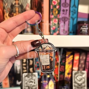May include: A clear plastic keychain charm shaped like a coffee cup with a brown liquid and ice cubes inside. The cup has a black label with the text "Order of 1 item in order: Dark Romance 1. Spice 2.  A grumpy good girl 3.  A hot stabby 4.  Twice morally grey".