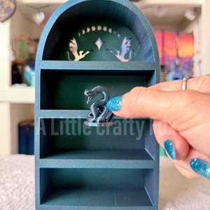 May include: A teal-colored, three-tiered shelf with an arched top. The top shelf has a decorative cutout design. A small, dark gray snake figurine sits on the middle shelf. The text "A Little Crafty Fox" is visible on the bottom shelf.