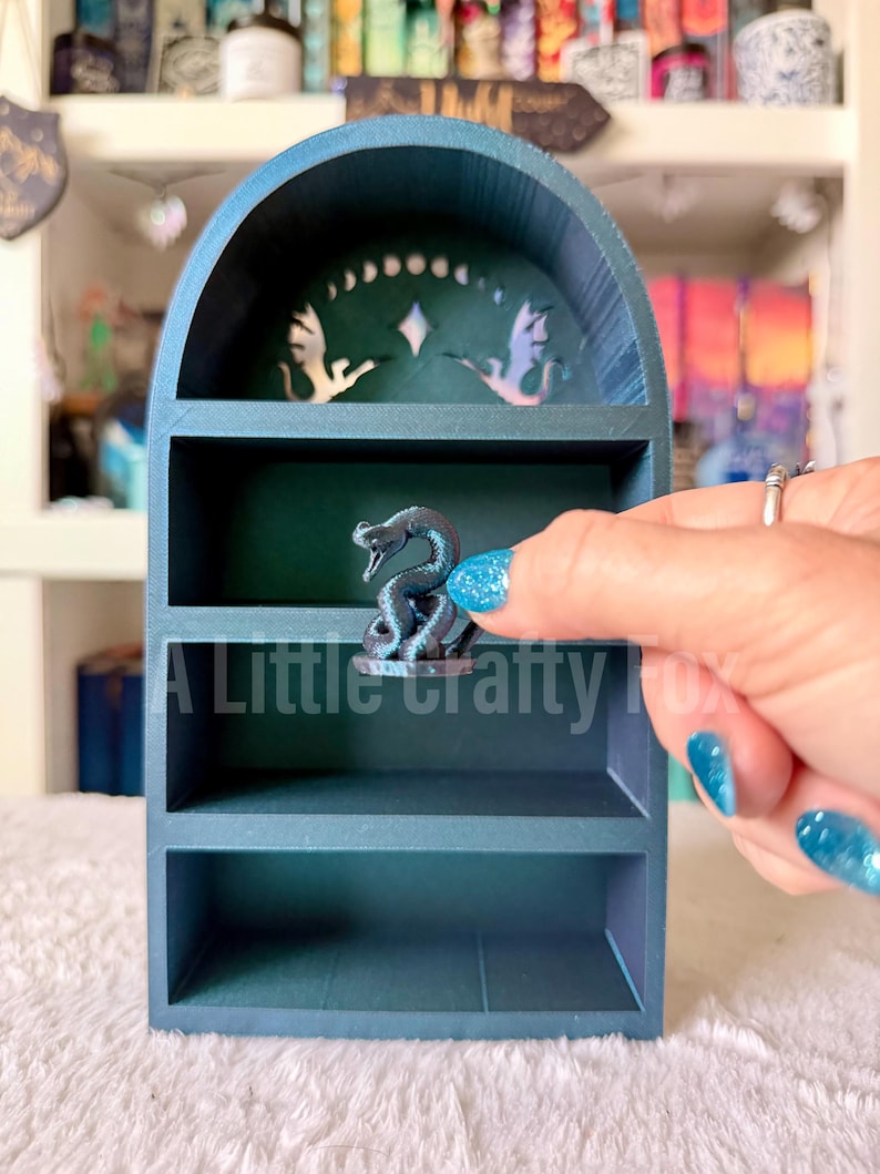May include: A teal-colored, three-tiered shelf with an arched top. The top shelf has a decorative cutout design. A small, dark gray snake figurine sits on the middle shelf. The text "A Little Crafty Fox" is visible on the bottom shelf.