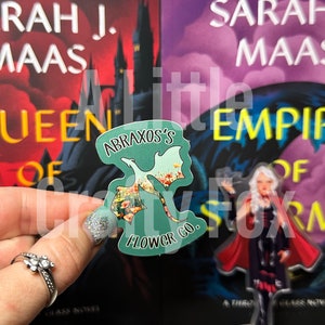 Puede incluir: Una pegatina verde con un diseño floral y una silueta de dragón. La pegatina dice "Abraxos's Flower Co."