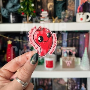 Peut inclure: Un autocollant de piment fantôme rouge avec un visage souriant et le texte "True Love is Eating Ghost Chili Peppers". L'autocollant a une finition brillante et est tenu par une main.