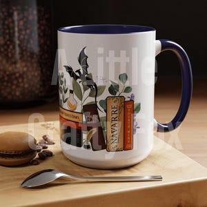 May include: White ceramic mug with a blue rim and handle. The mug features a black dragon, green leaves, and a stack of books with titles like "Navarre" and "Fourth Wing".