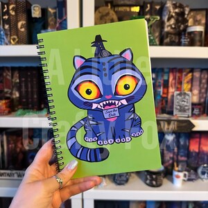 以下が含まれることがあります： 鮮やかな緑色の表紙に漫画の猫のイラストが描かれた、スパイラル綴じのノート。猫は青色で、黄色い目と黒い帽子をしています。「Save the Date」の文字がバナーに書かれています。ノートは手で持たれています。