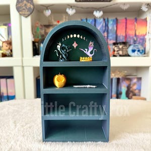 May include: A teal-colored, three-tiered shelf unit with an arched top. The top shelf features a dragon and a winged creature. The middle shelf holds a golden apple. The bottom shelf has the text "Little Crafty Fox".