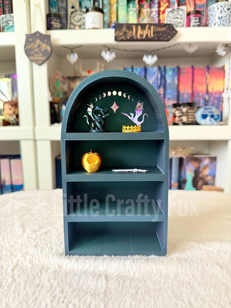 May include: A teal-colored, three-tiered shelf unit with an arched top. The top shelf features a dragon and a winged creature. The middle shelf holds a golden apple. The bottom shelf has the text "Little Crafty Fox".