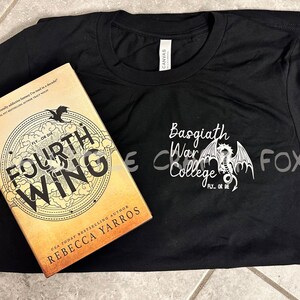 Pu&ograve; includere: Una maglietta nera con la scritta "Basgiath War College" e un disegno di drago. Accanto alla maglietta c'&egrave; un libro intitolato "Fourth Wing" di Rebecca Yarros, con un design circolare e le parole "Fly or Die".