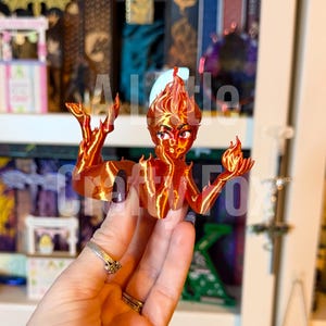 May include: A 3D-printed figurine of a fiery mermaid with orange and red hues. The mermaid has flames for hair and is holding up her hands. The figurine is held in a hand, with a blurred background of shelves and other decorative items.