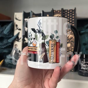 May include: White ceramic mug with a black handle. The mug features a black and white illustration of a dragon, a bottle, a quill pen, and three books. The books have the titles "NAVARRE", "FOURTH WING", and "DRAGON RIDER'S GODDEN".