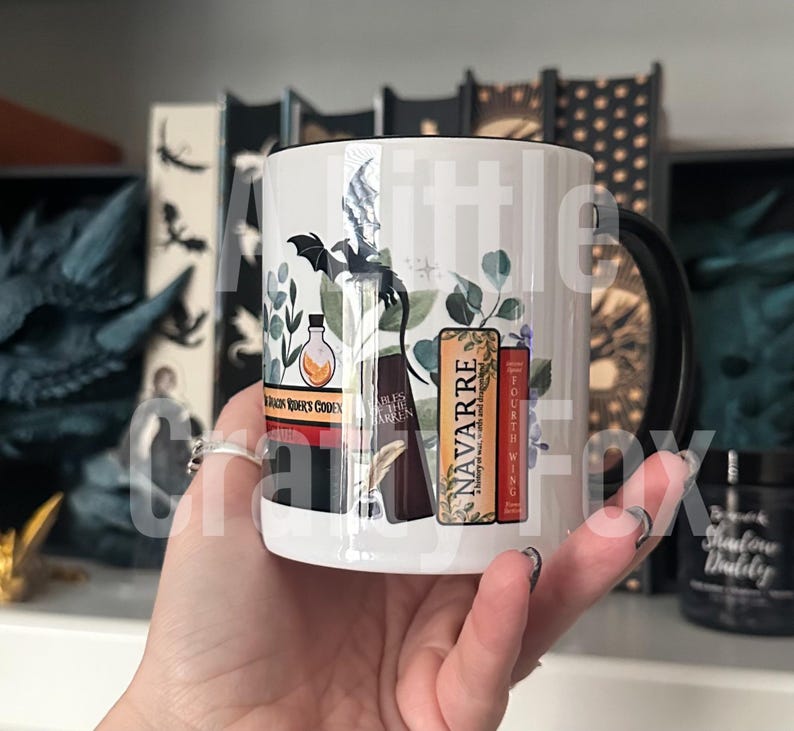 May include: White ceramic mug with a black handle. The mug features a black and white illustration of a dragon, a bottle, a quill pen, and three books. The books have the titles "NAVARRE", "FOURTH WING", and "DRAGON RIDER'S GODDEN".
