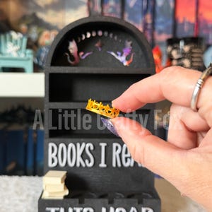 May include: A black miniature bookshelf with the text "BOOKS I READ THIS YEAR." The shelf has a curved top with decorative cutouts. A small gold-colored crown is held in front of the shelf. A stack of miniature books sits on the shelf.