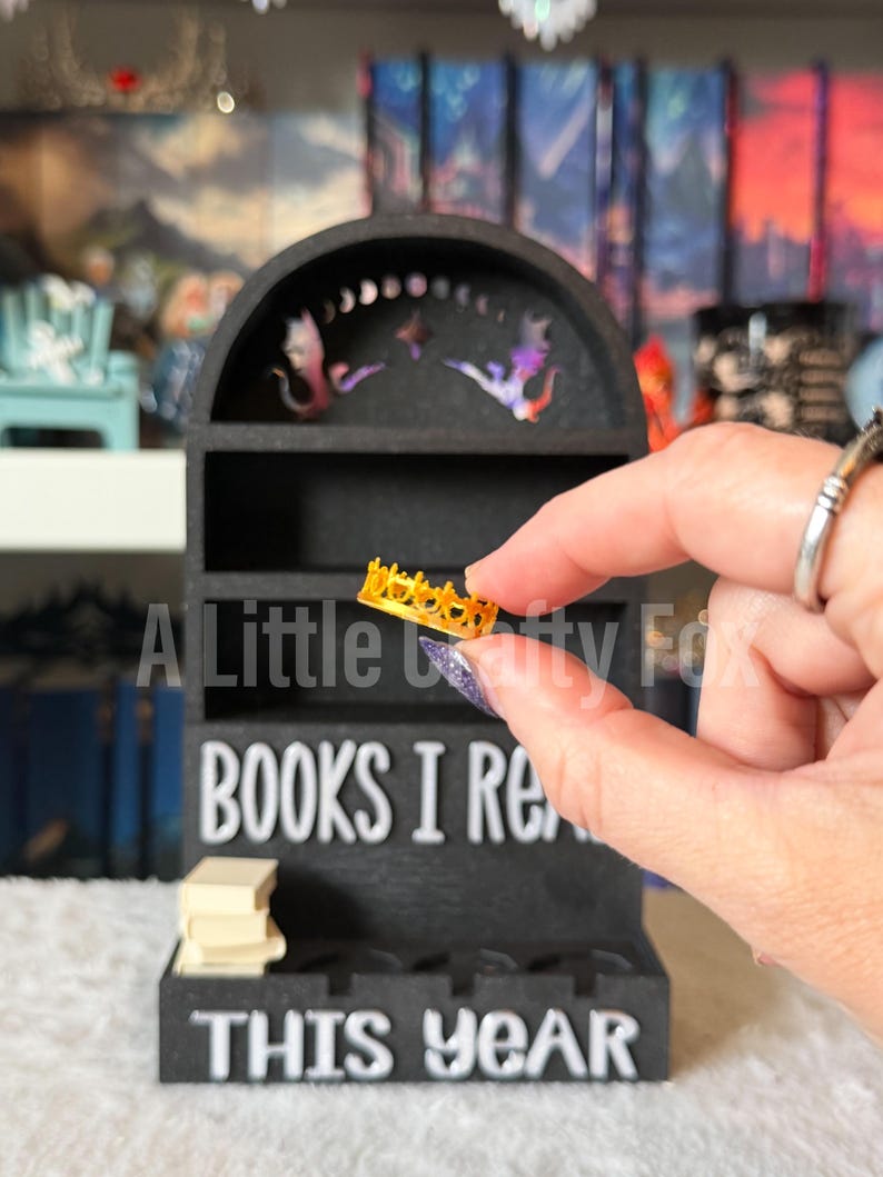 May include: A black miniature bookshelf with the text "BOOKS I READ THIS YEAR." The shelf has a curved top with decorative cutouts. A small gold-colored crown is held in front of the shelf. A stack of miniature books sits on the shelf.