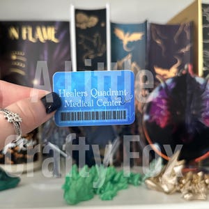 Peut inclure: Une carte bleue avec le texte "Healers Quadrant Medical Center" et un code-barres. La carte est tenue par une main avec une bague en argent.
