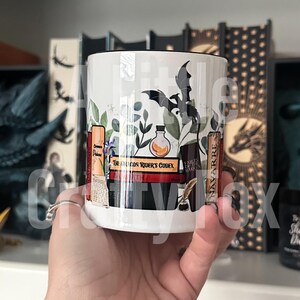 May include: White ceramic mug with a black rim and handle. The mug features a whimsical design of a bat, books, and a potion bottle with a green leafy background. The text on the books reads "Dragon Rider's Codex" and "Ember Earth Hark".