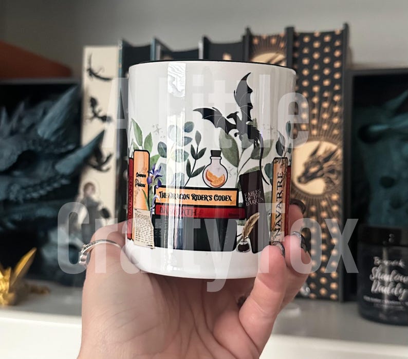 May include: White ceramic mug with a black rim and handle. The mug features a whimsical design of a bat, books, and a potion bottle with a green leafy background. The text on the books reads "Dragon Rider's Codex" and "Ember Earth Hark".