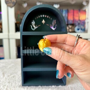 May include: A teal-colored, arched shelf with two shelves. The top of the shelf has a decorative design with two birds and a moon phase. A small, yellow, apple-shaped object is held in front of the shelf.