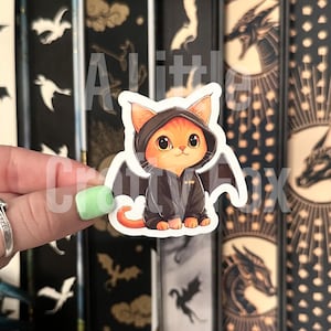 May include: A sticker featuring an orange cat wearing a black hooded jacket with bat wings. The cat has large, expressive eyes and is sitting upright. The sticker has the text "A Little Crafty Box".