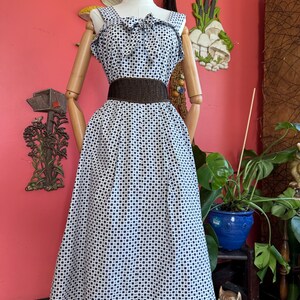 May include: A sleeveless white dress featuring a pattern of small dark blue polka dots, a square neckline with a decorative bow detail at the center, and a wide dark brown textured belt cinched at the waist, displayed on a mannequin.