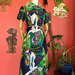 May include: A knee-length dress featuring a vibrant botanical print with large green, purple, and white leaves on a dark background. The dress has short sleeves, a pointed collar, a front zipper closure, and a thin beige belt at the waist.