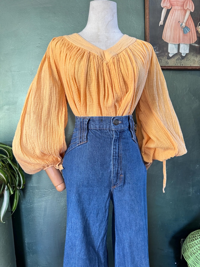 May include: A vintage ensemble featuring a peach-colored blouse with a V-neck and long, gathered sleeves. The blouse is paired with high-waisted, wide-leg blue denim jeans. The outfit is displayed on a mannequin.