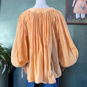 May include: A peach-colored blouse with long, voluminous sleeves and a gathered neckline. The fabric has a textured appearance. The blouse is displayed on a mannequin, paired with blue jeans. The style is reminiscent of vintage or bohemian fashion.