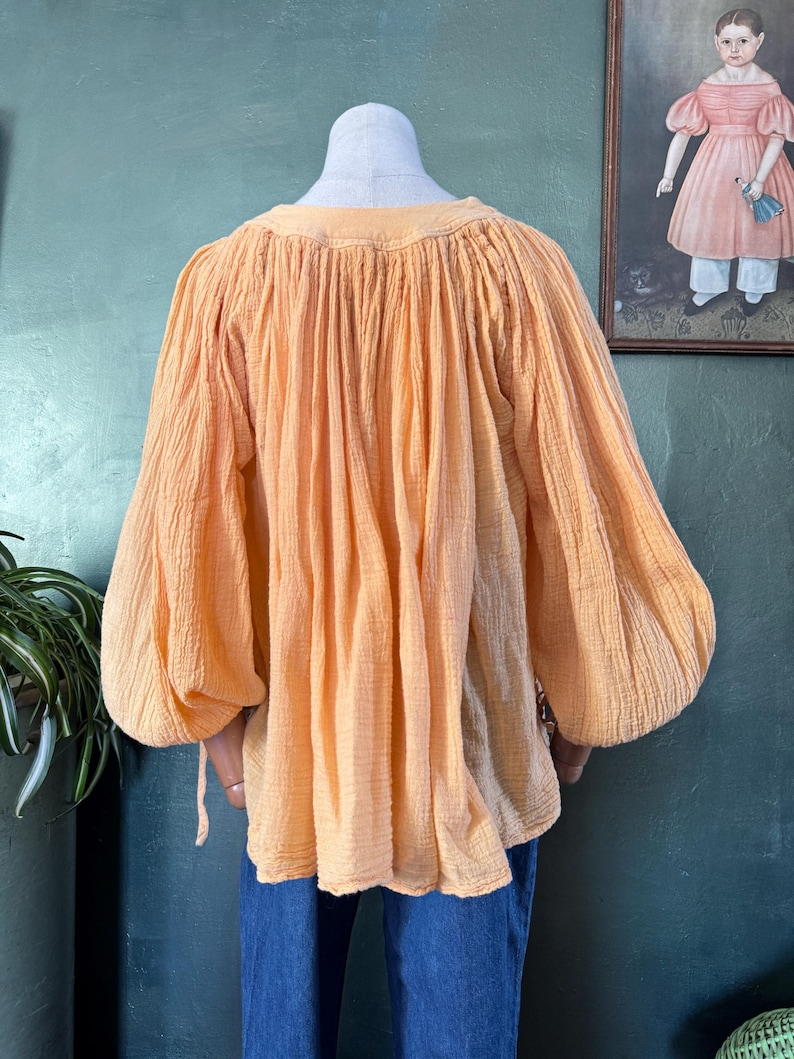 May include: A peach-colored blouse with long, voluminous sleeves and a gathered neckline. The fabric has a textured appearance. The blouse is displayed on a mannequin, paired with blue jeans. The style is reminiscent of vintage or bohemian fashion.